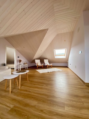 Een zolderkamer met houten vloeren, dakraam, een witte bank, twee stoelen, een klein tafeltje en een decoratieve mand.