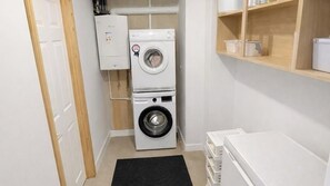 A compact laundry room with a front-loading washer and dryer, a small shelf with containers, and a door leading to another room. - NEW!4BR Ideal for Contractors, Driveway, Wifi, M40 (Heathcote)