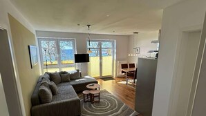 A modern living room with a sectional sofa, a dining area with a table and chairs, and a kitchen area with a refrigerator. - Ferienwohnung in Orth auf Fehmarn (Fehmarn)