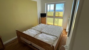A bedroom with a bed, wooden drawers, a desk with a computer, and a window with blinds. - Ferienwohnung in Orth auf Fehmarn (Fehmarn)