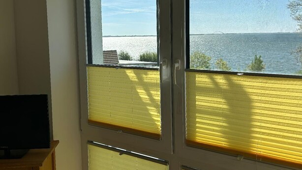 A room with a large window overlooking a body of water, a television on a stand, and a door. - Ferienwohnung in Orth auf Fehmarn (Fehmarn)