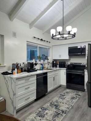 A modern kitchen with white cabinetry, a black stove, a microwave, and a dishwasher. There is a window with blue blinds, a chandelier, and a patterned rug on the floor. - Perfect 2-bedroom condo & yard. Newport Beach, Balboa Pier, Fun Zone, w Parking (Newport Beach)