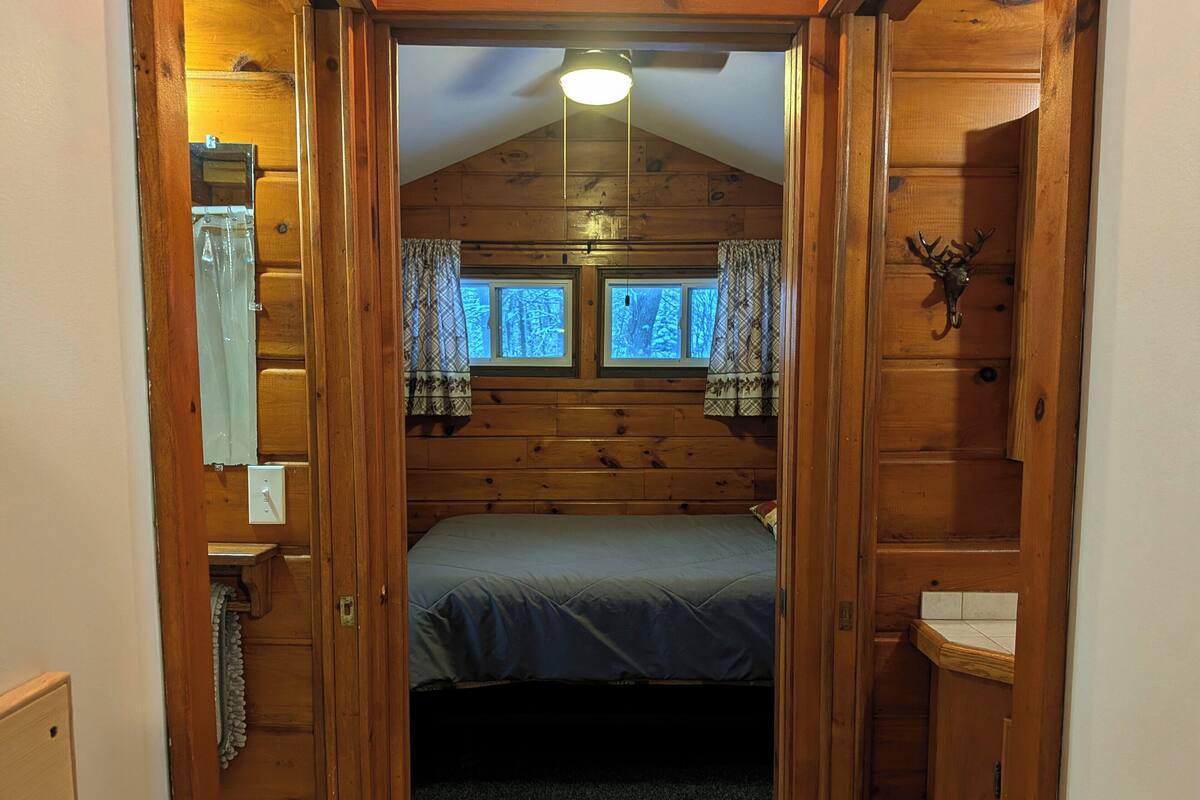 A wooden cabin interior with a bed, two windows with curtains, a ceiling light, and a wall-mounted light fixture.