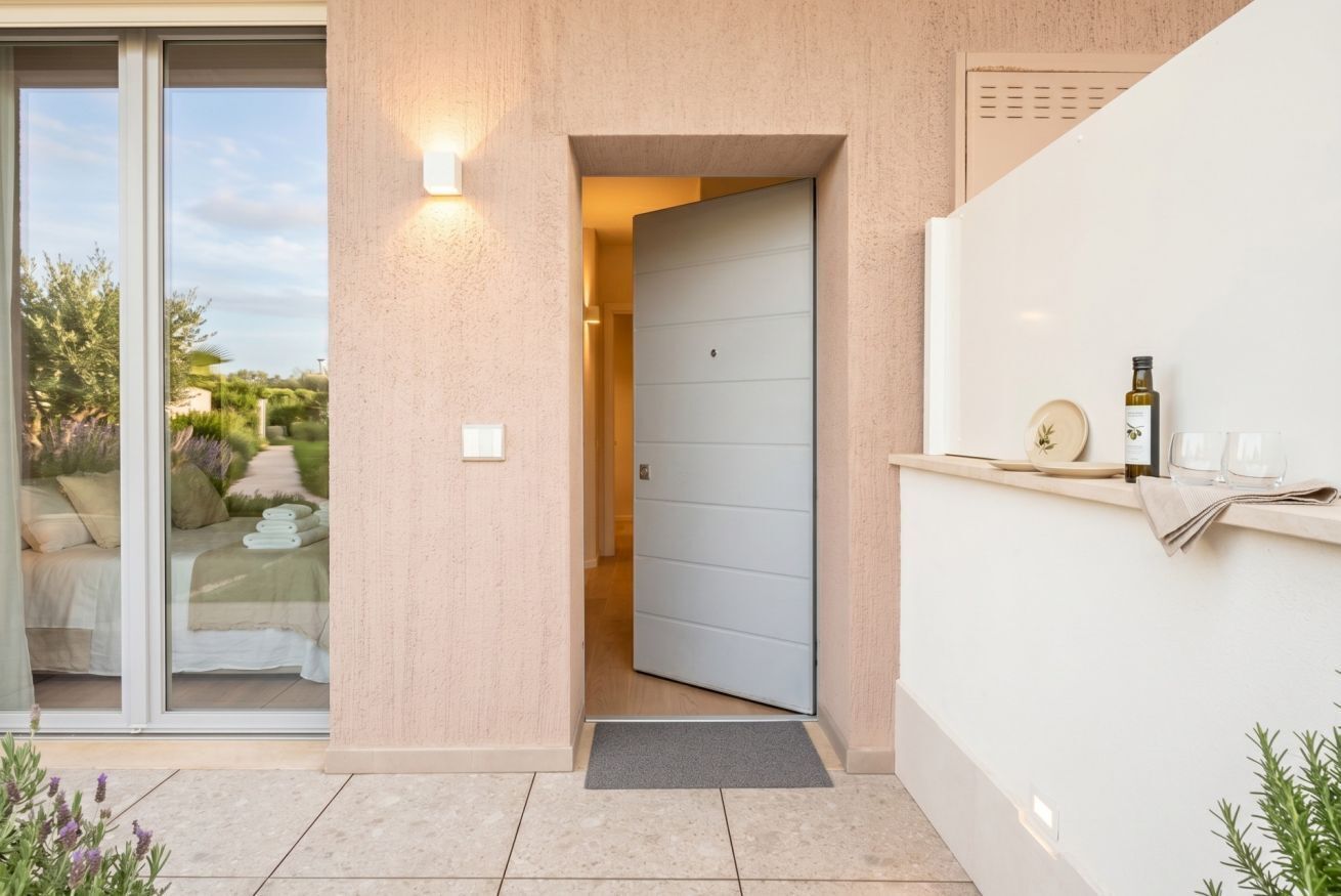 Ingresso di un edificio moderno con porta scorrevole in vetro che conduce a una camera da letto, una porta azzurra e un piccolo spazio all'aperto con una bottiglia e dei bicchieri.