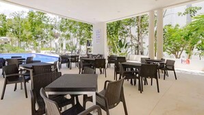 Un patio al aire libre con mesas y sillas, una piscina y vegetación circundante.