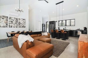 A modern living room with a sectional sofa, dining table, and chairs, a kitchen with a refrigerator and bar stools, and abstract art on the walls.