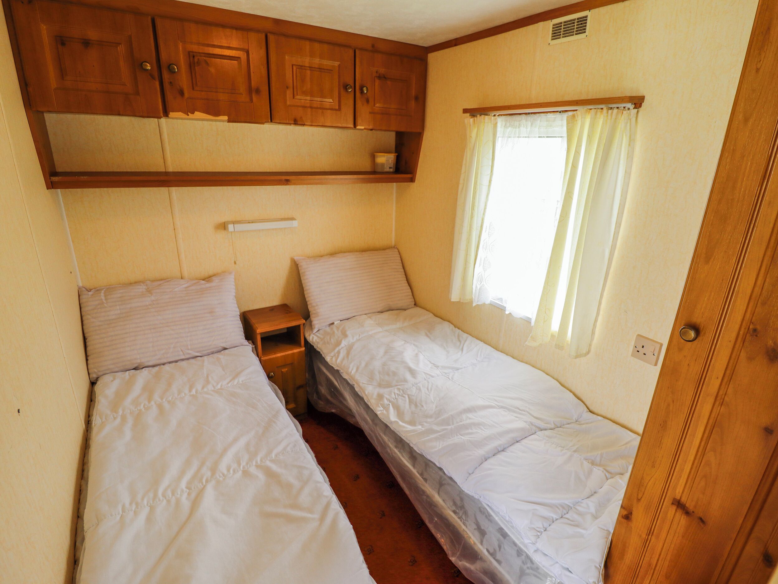 Un dormitorio de autocaravana compacto con dos camas, armarios de madera arriba, una ventana con cortinas y un enchufe.