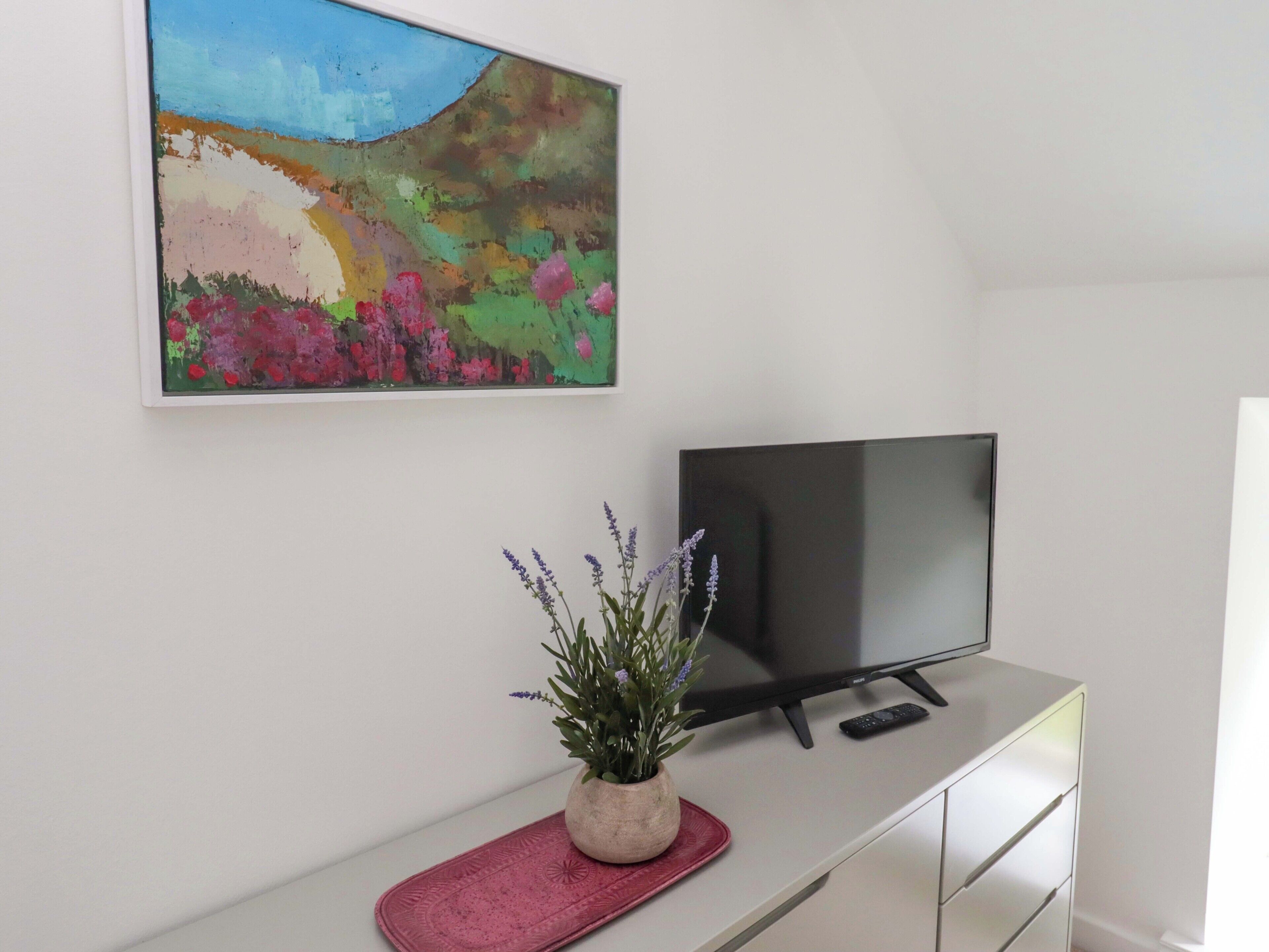 Ein Zimmer mit einem Flachbildfernseher auf einer weißen Kommode, einem farbenfrohen abstrakten Gemälde an der Wand und einer Topfpflanze auf einer roten Matte.