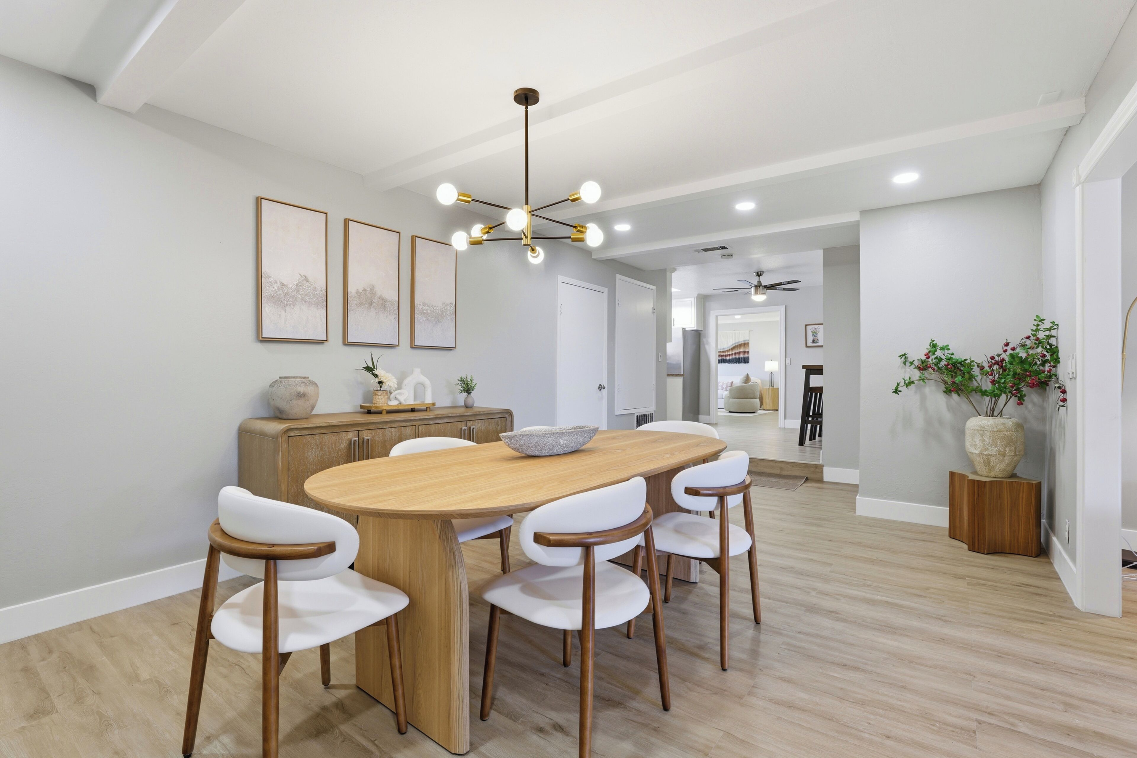 Un comedor con una mesa de madera, sillas blancas, un artefacto de iluminación central y una cocina al fondo.