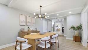 Uma sala de jantar com uma mesa de madeira, cadeiras brancas, um lustre central e uma cozinha ao fundo.