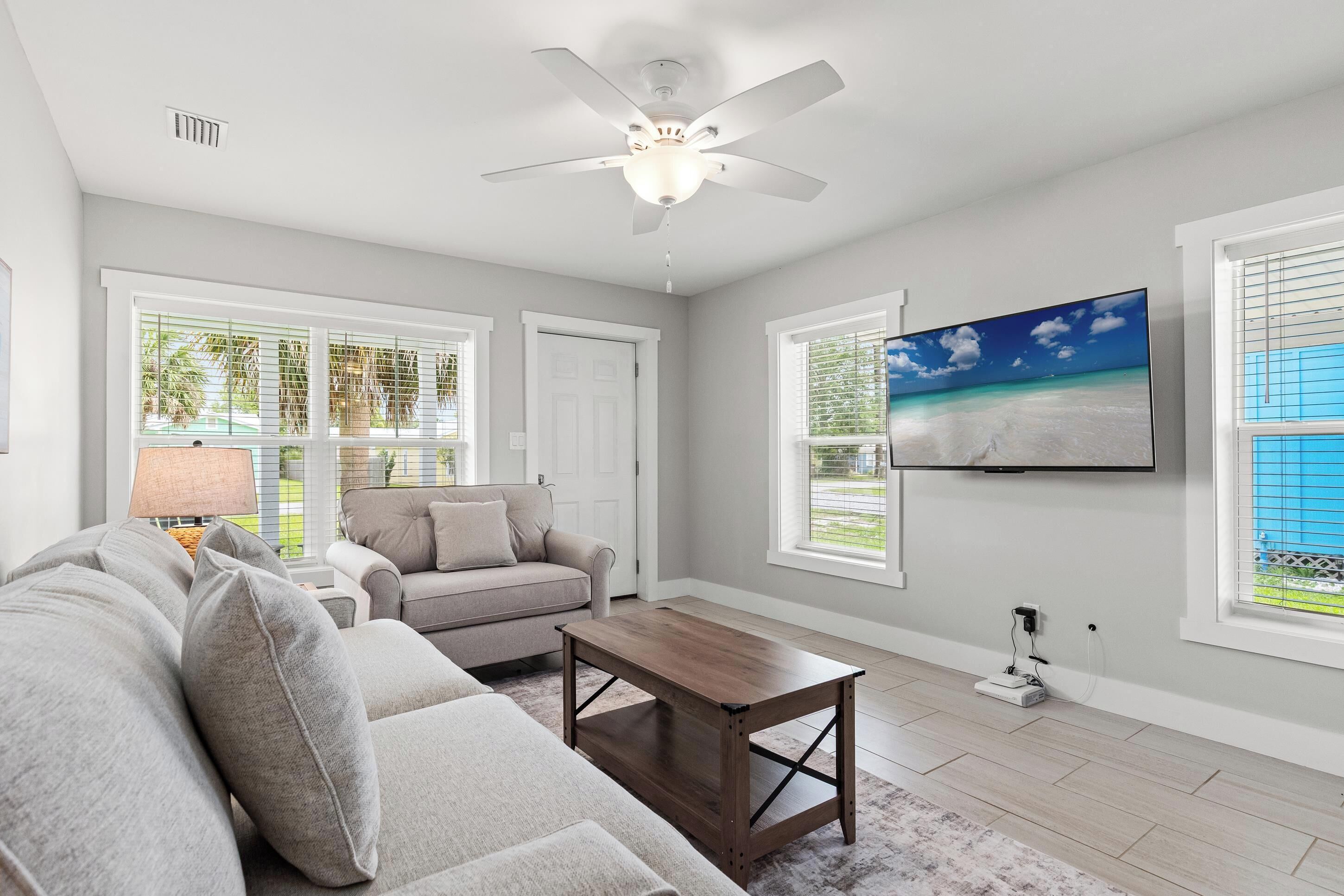 A living room with a ceiling fan, a TV mounted on the wall, a sofa, a coffee table, and large windows with blinds.