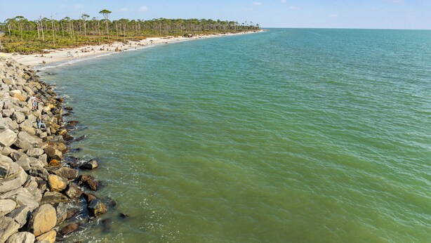 A beach with a rocky shoreline, clear turquoise water, and a line of trees in the background. - Beach House - St Joe Retreat (Port St. Joe)