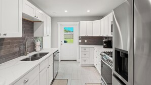 A modern kitchen with white cabinets, stainless steel appliances, and a view of a green landscape through a door. - Beach House - St Joe Retreat (Port St. Joe)