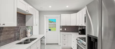 A modern kitchen with white cabinets, stainless steel appliances, and a view of a green landscape through a door.