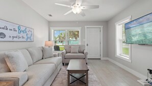 A living room with a sofa, a coffee table, a ceiling fan, a TV, and a window with blinds. - Beach House - St Joe Retreat (Port St. Joe)