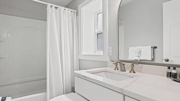 Baño con ducha, inodoro, lavabo de dos grifos y espejo.