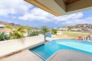 A swimming pool with steps leading into it, surrounded by palm trees and a view of a golf course and buildings in the background. - NEW | 4BR Villa with Private Pool at Blue Bay (Sint Michiel)