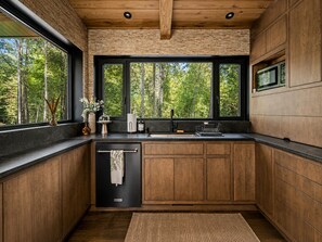 A modern kitchen with a built-in dishwasher, wooden cabinets, and a large window offering a view of a forest. - Luxury Treehouse Retreat | Bordering Gorges (Brevard)