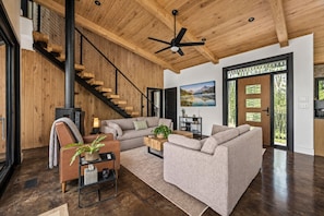A modern living room with a wooden staircase, a fireplace, and a large window offering a view of the outdoors. - Luxury Treehouse Retreat | Bordering Gorges (Brevard)