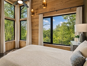 A bedroom with a large bed, wooden walls, and a view of trees through large windows. - Luxury Treehouse Retreat | Bordering Gorges (Brevard)