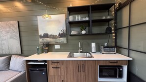 A compact kitchen with a dishwasher, microwave, and a small refrigerator. It features a wooden cabinet with a built-in sink and a hanging pendant light. - The Persimmon Tiny House at Cyprus Trail (Rockbridge)