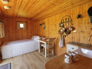 A wooden cabin with a bed, a dining table, chairs, a kitchen area with a sink, and a hanging pot rack.