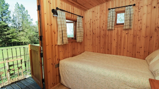A wooden cabin bedroom with a bed, two windows with curtains, and a balcony. - Bills Shepherds Hut (Brecon)