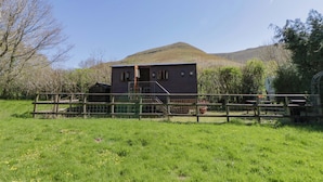 A rustic wooden building with a balcony, surrounded by a fenced area and a grassy field, with a mountain in the background. - Bills Shepherds Hut (Brecon)