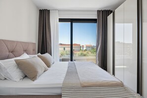 A bedroom with a bed, curtains, and a view of buildings through a large window. - Beautiful 3-bedroom apartment with AC in charming Kaštel Stari (Kaštel Stari)