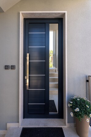 A modern black door with a vertical handle, a small potted plant with white flowers, and a set of stairs leading to an upper level. - Beautiful 3-bedroom apartment with AC in charming Kaštel Stari (Kaštel Stari)