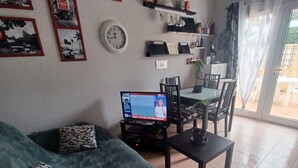 Un saló amb un sofà verd, almofadons estampats, una televisió sobre un suport, una zona de menjador amb cadires i una taula, i un rellotge de paret.