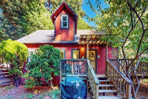 A red house with a porch, a covered deck, and a staircase leading to the entrance. - Cazadero 2BR creekside cottage — fenced yard, dogs welcome (Cazadero)