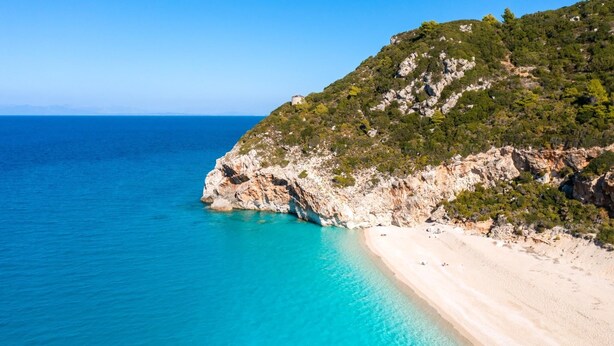 Ein Küstenstrand mit klarem türkisblauem Wasser, einem Sandufer und einem felsigen Hang mit spärlicher Vegetation.