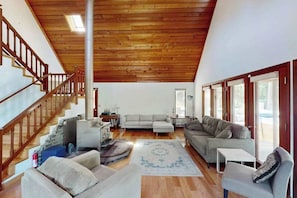 A spacious living area with a wooden ceiling, a staircase, a gray sofa, a white armchair, and a patterned rug.