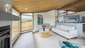 A modern living room with a fireplace, a sofa, a round wicker coffee table, and a kitchen area with a view of the outdoors. - Architect-Designed Bodega Bay Home w/ Ocean Views & Hot Tub - 2 King Suites (Bodega Bay)
