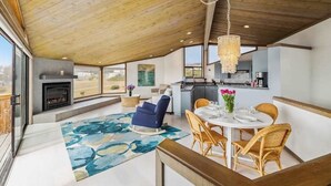 A spacious living area with a fireplace, dining table, and chairs, a kitchen in the background, and a large window offering a view. - Architect-Designed Bodega Bay Home w/ Ocean Views & Hot Tub - 2 King Suites (Bodega Bay)
