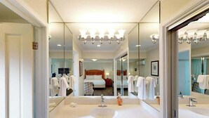 Uma casa de banho de hotel com duas pias, um espelho grande e uma cama visível através do espelho.