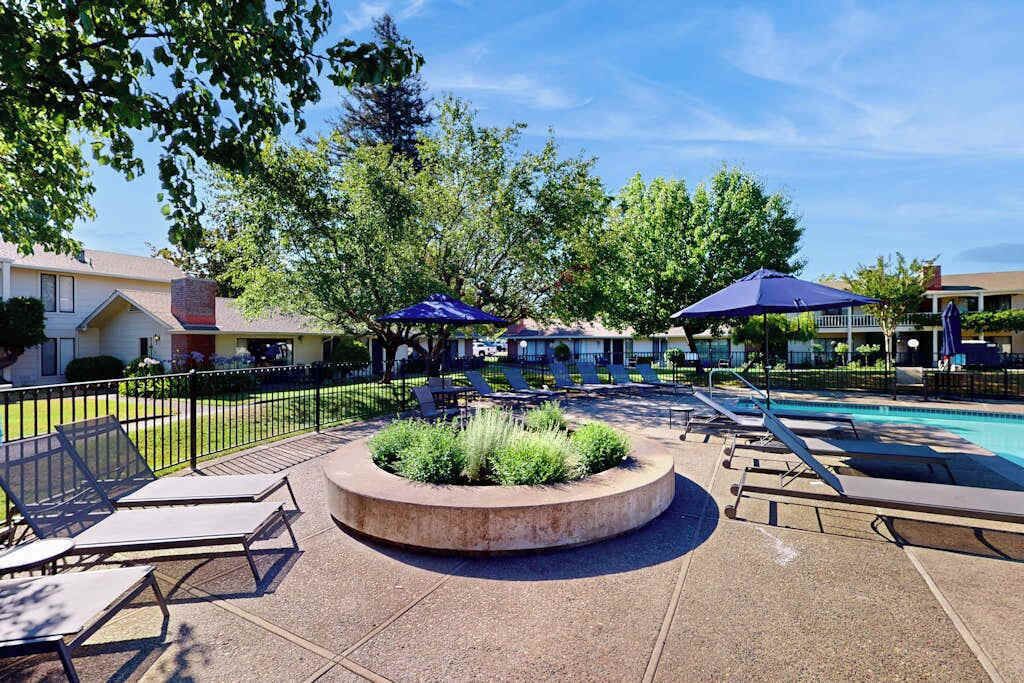 A pool area with lounge chairs and umbrellas, surrounded by trees and a building.