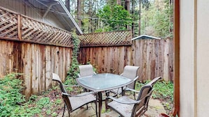 A wooden deck with a glass table and chairs, surrounded by a wooden fence and trees.
