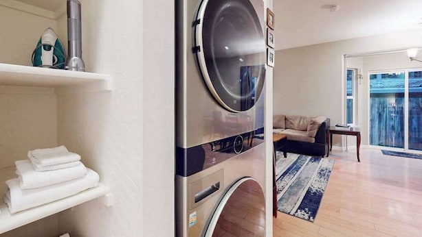 A modern laundry room with a built-in washer and dryer, a shelf with folded towels, and a view of a living area with a sofa and a large window.
