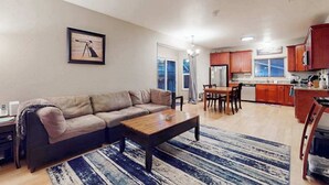 A living room with a sofa, coffee table, and a rug. There is a dining area with a table and chairs, and a kitchen with cabinets and appliances.