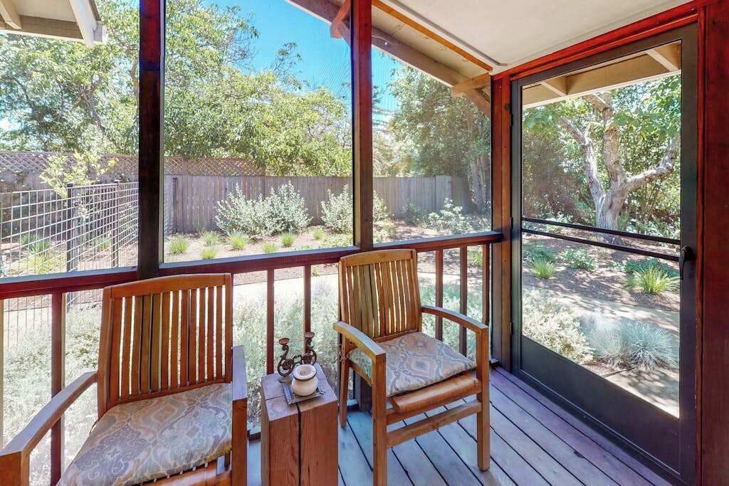 A covered outdoor area with two wooden chairs and a small table, a metal railing, and a view of a backyard with trees and a fence.
