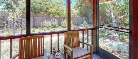 A covered outdoor area with two wooden chairs and a small table, a metal railing, and a view of a backyard with trees and a fence.