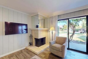 Ein Wohnzimmer mit Kamin, Flachbildfernseher, einer Couch und Blick durch eine Glastür.