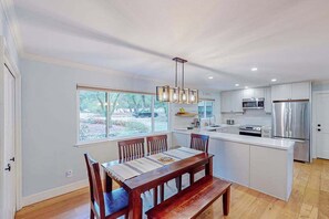 A modern kitchen with white cabinets, stainless steel appliances, a wooden dining table, and a large window.