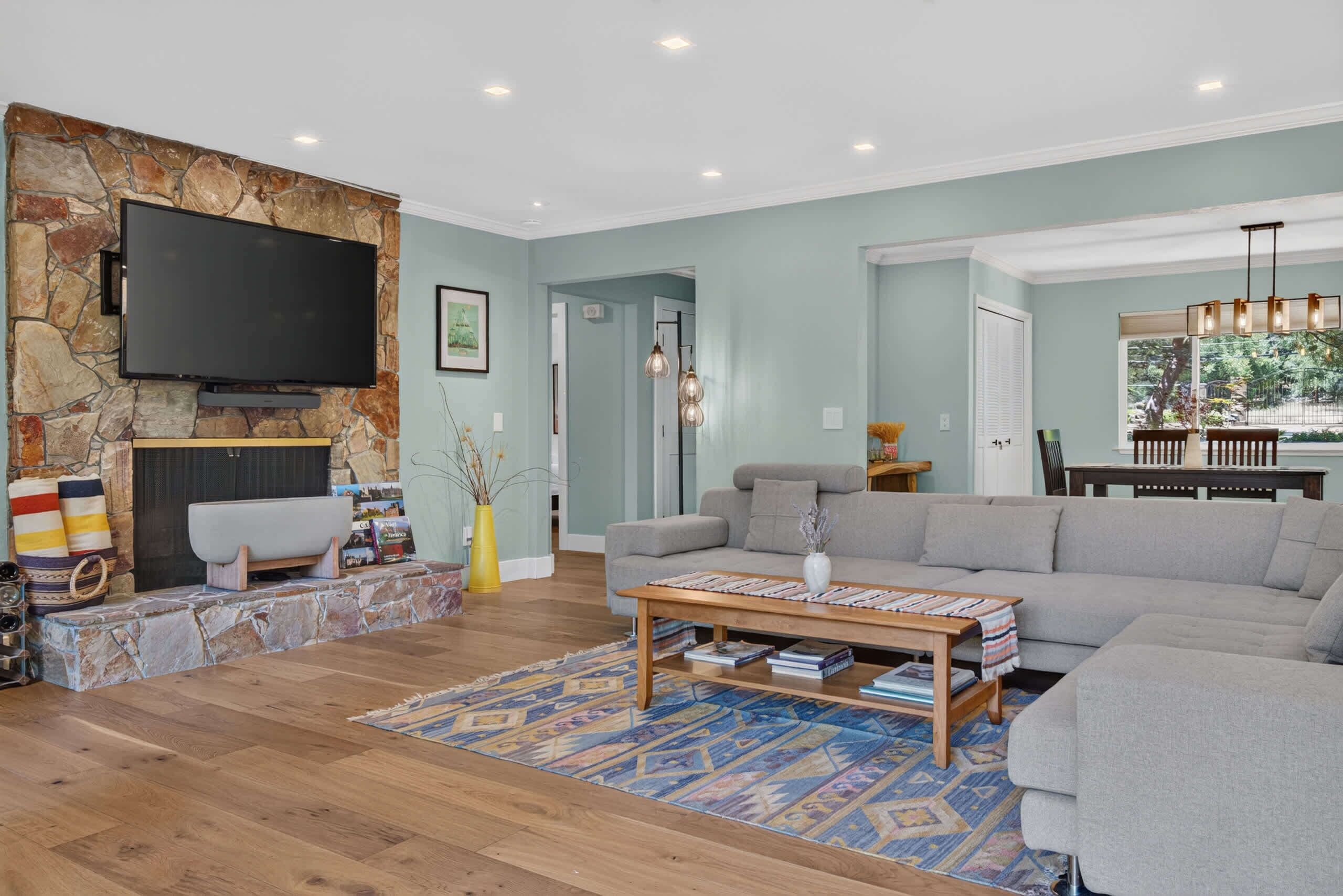 A living room with a stone fireplace, a flat-screen TV, a grey sofa, a coffee table, and a patterned rug.