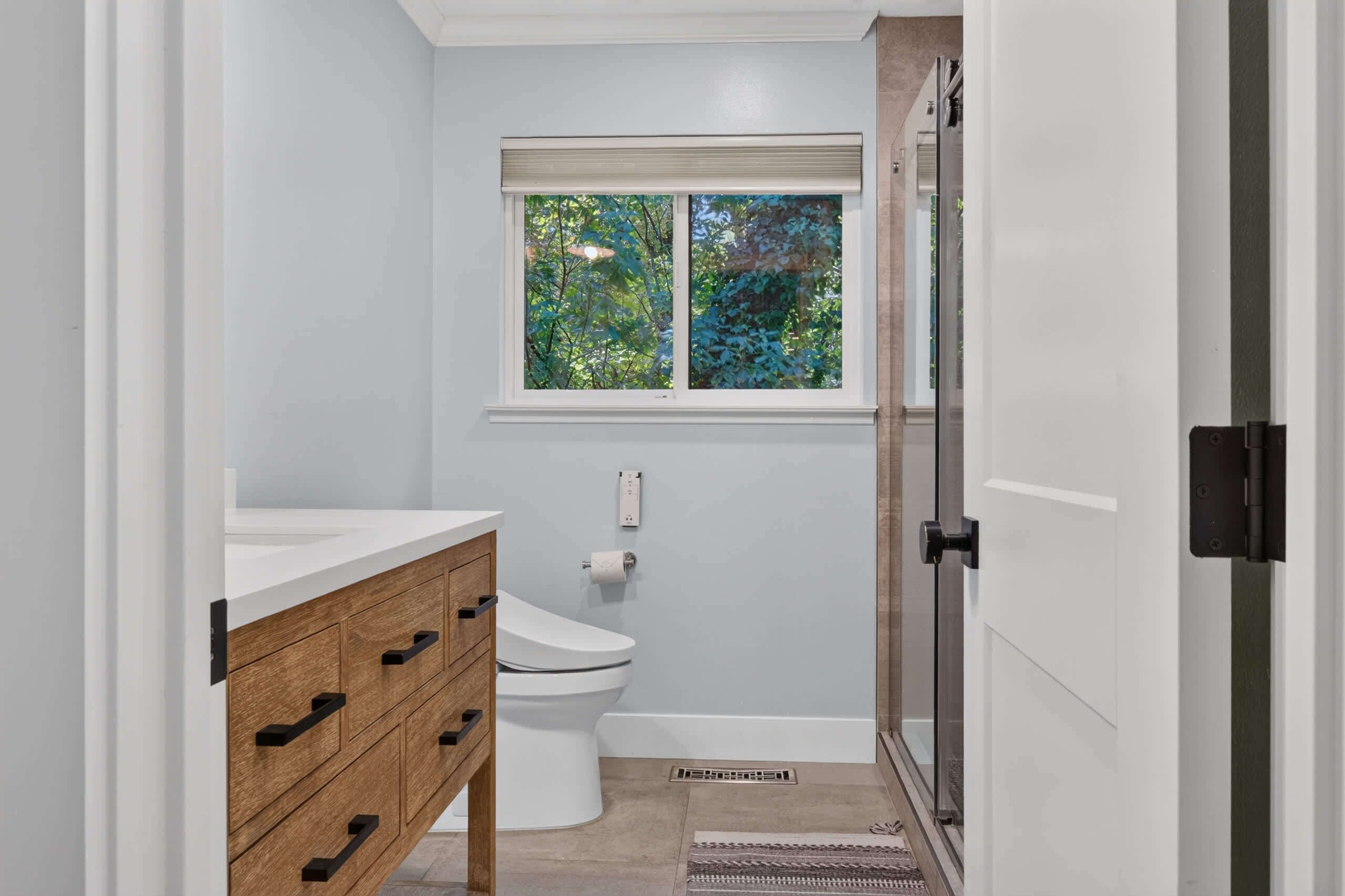 A bathroom with a toilet, a wooden vanity, a window with a view of trees, and a shower area.