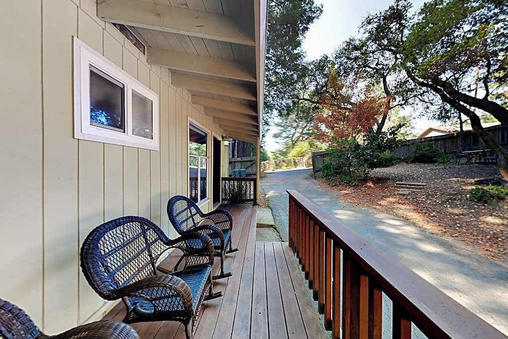 Un porche cubierto con mobiliario exterior, una terraza de madera y vistas a un jardín ajardinado con árboles y una valla.