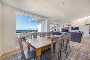 A dining area with a view of the sea, a kitchen with white cabinets, and a living area with a sofa and TV.