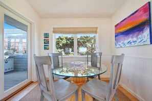 Un comedor con una mesa de vidrio y cuatro sillas, un ventanal con vistas a árboles, y un cuadro colorido en la pared.